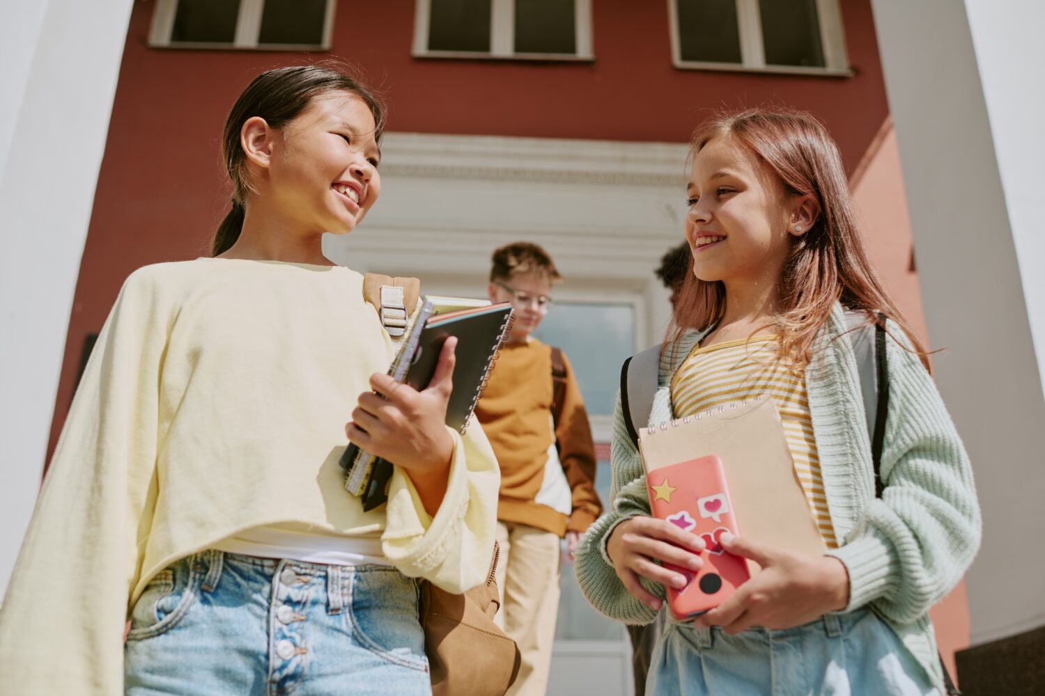 Two preteen girls, one Asian and one Caucasian, smiling and talking while holding notebooks and walking down steps outside school building, boy with backpack following behind