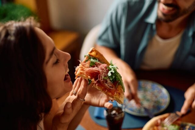 Close up of woman with eyes closed tasting pizza while eating with her boyfriend in restaurant.