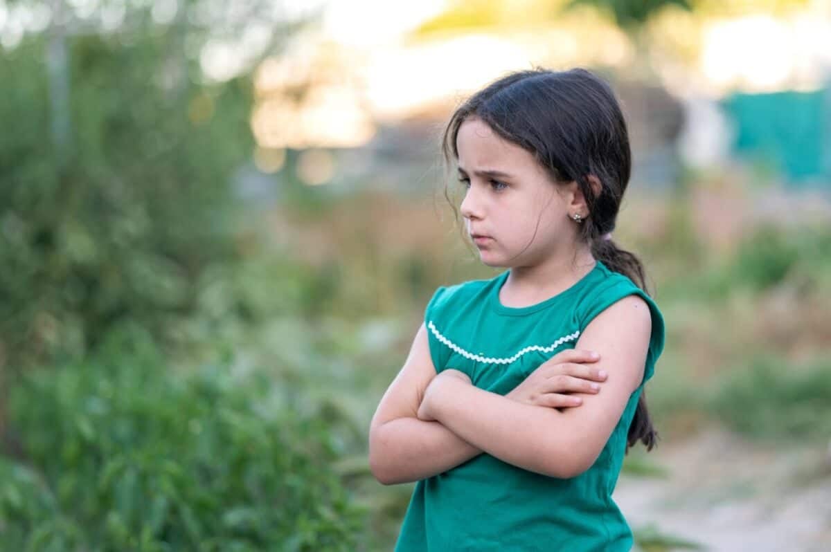 Upset child with a pouting expression standing outside with crossed arms