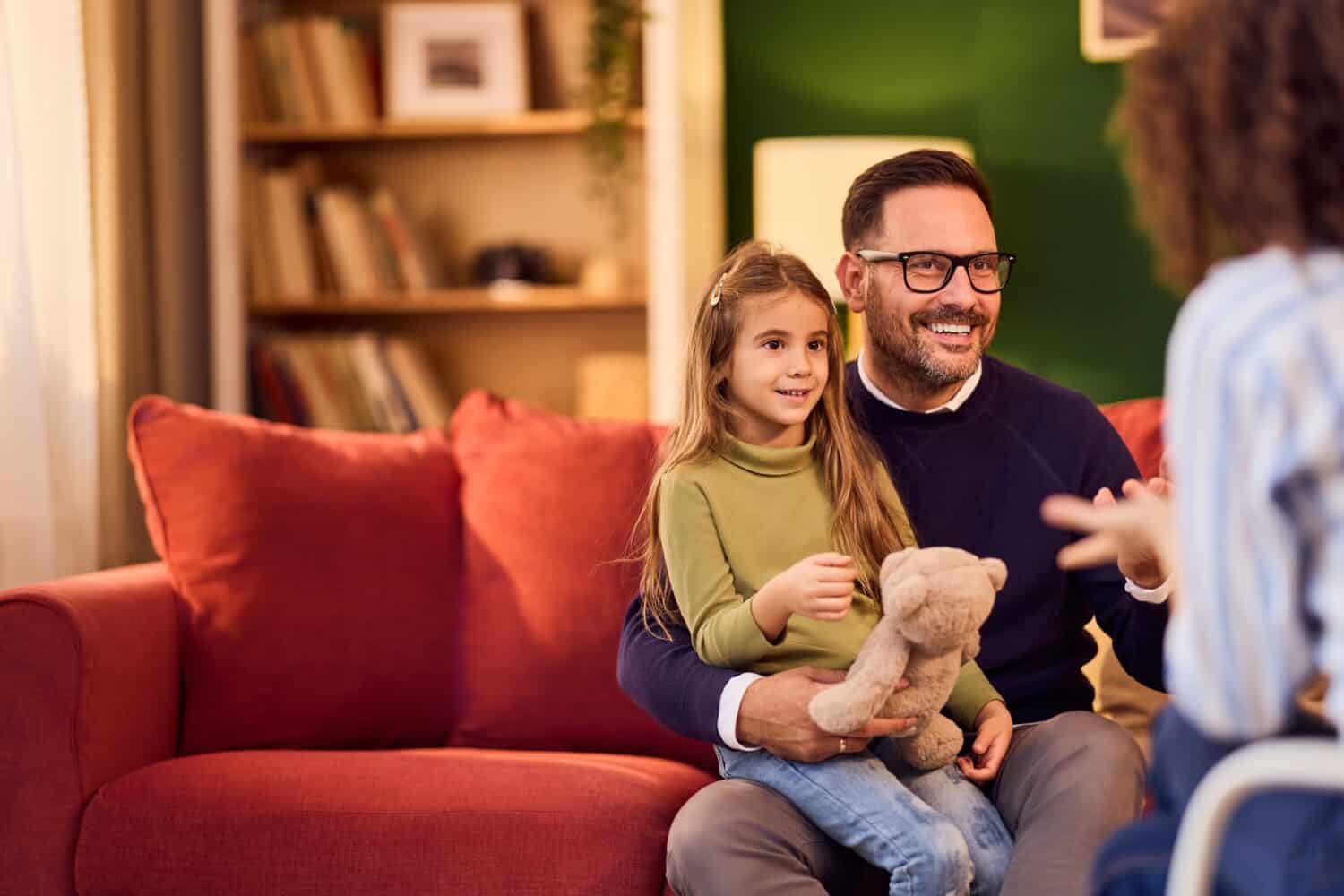 A father and daughter sit on a red sofa during a psychologist visit, smiling and relaxed.