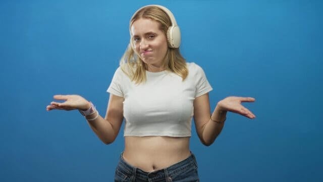 Woman with headphones touching one earcup and shrugging, midriff exposed in studio; bemusement doubt.
