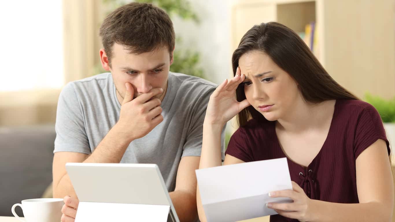A young man in a gray t-shirt looks distressed, hand covering his mouth, while looking at a white tablet. Beside him, a young woman in a maroon top holds and reads a white letter, her face showing worry and her hand resting on her forehead. They appear to be in a living room or home office.