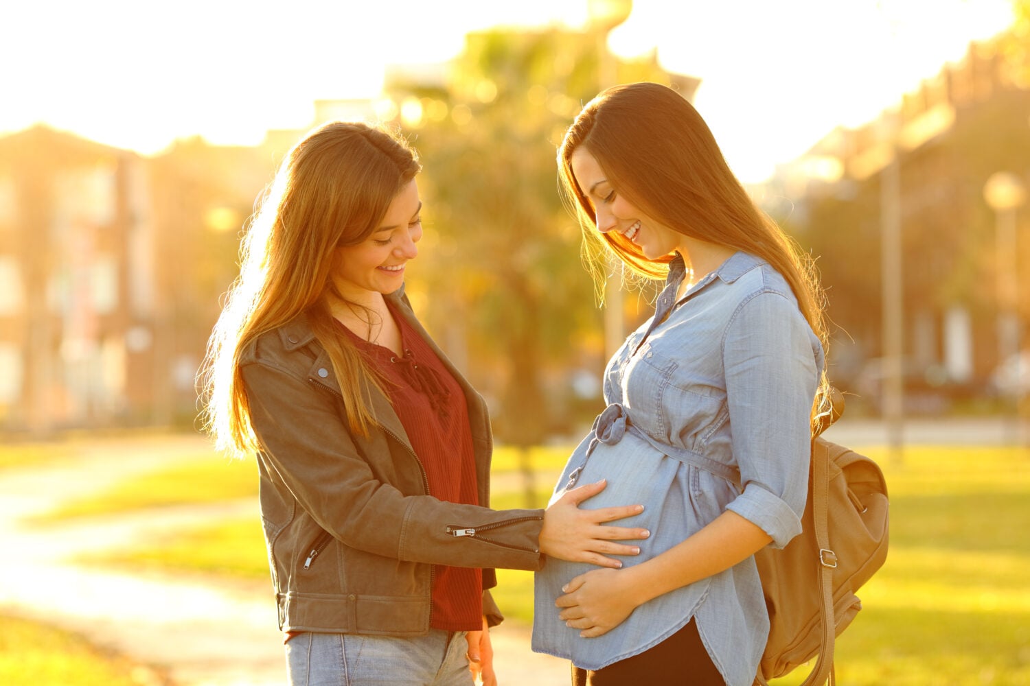 Happy pregnant woman showing belly to a friend in a park at sunset