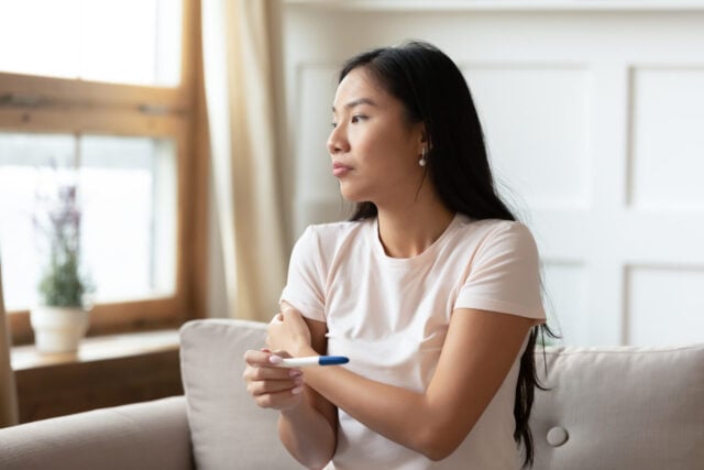 Pensive young Asian woman sit on couch hold ovulation pregnancy test wait for positive negative result, Vietnamese millennial girl have health problems, IVF treatment, fertility infertility concept