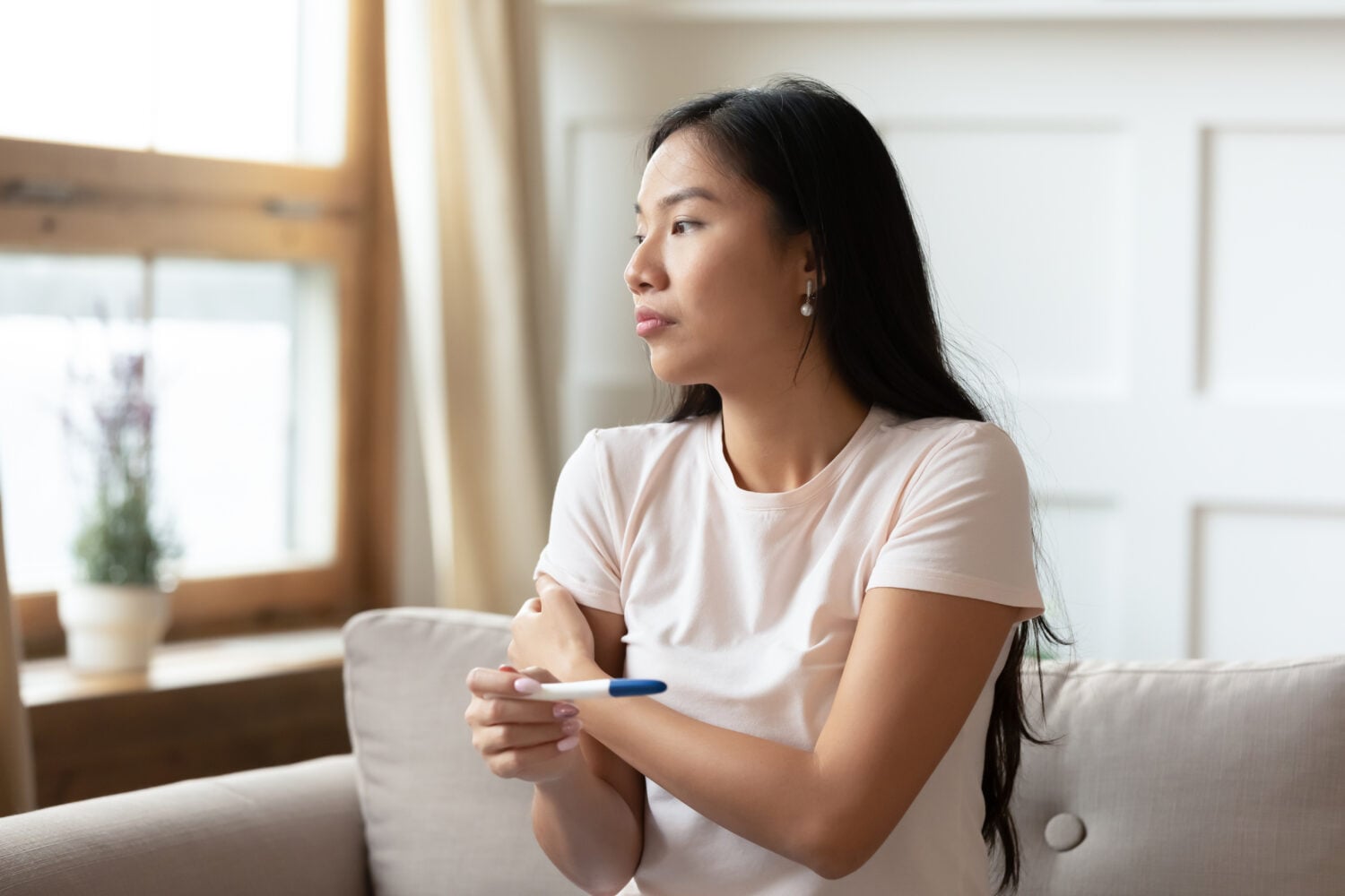 Pensive young Asian woman sit on couch hold ovulation pregnancy test wait for positive negative result, Vietnamese millennial girl have health problems, IVF treatment, fertility infertility concept
