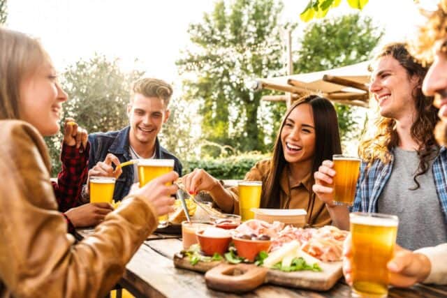 Group of multi ethnic friends having backyard dinner party together - Diverse young people sitting at bar table toasting beer glasses in brewery pub garden - Happy hour, lunch break and youth concept