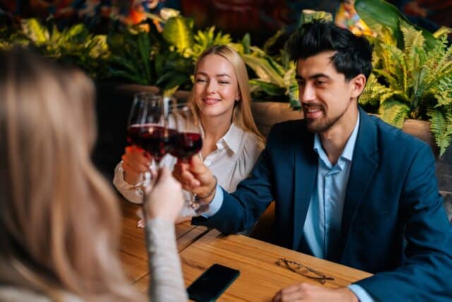 View from back of unrecognizable female clinking glass of red wine with smiling beautiful man and woman sitting at table in fancy restaurant at evening. Happy three friends enjoying nice dinner.