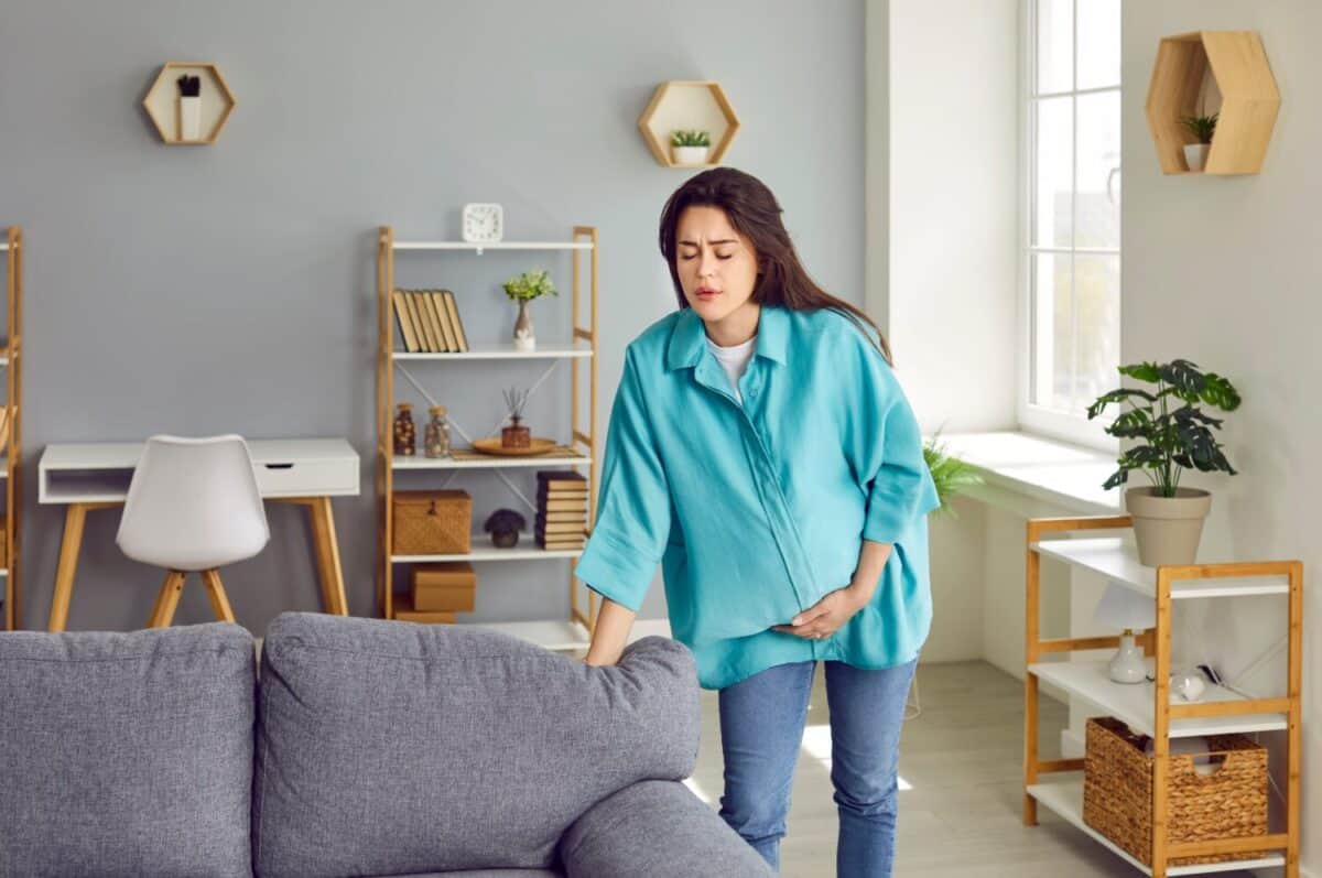 Pregnant woman feels prenatal cramps, discomfort and painful baby bumps or has acute pain as labor contractions start. Woman stands in living room, leans on couch and grabs her belly Pregnancy concept