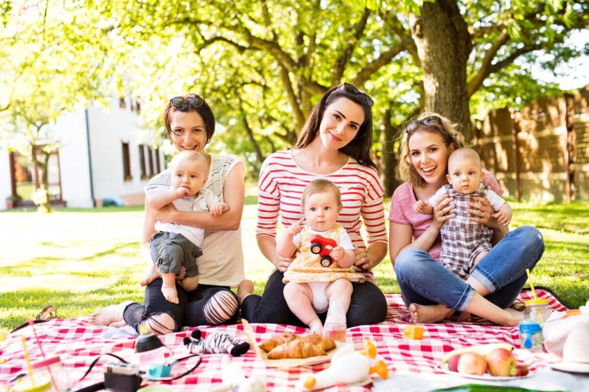 Mothers and babies enjoying group picnic outdoor in park, sitting on picnic blanket and preparing food and drinks.