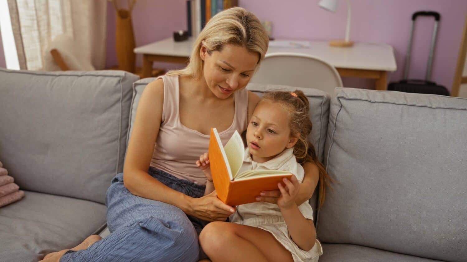 Woman reading book to daughter on couch in living room with cozy interior setting