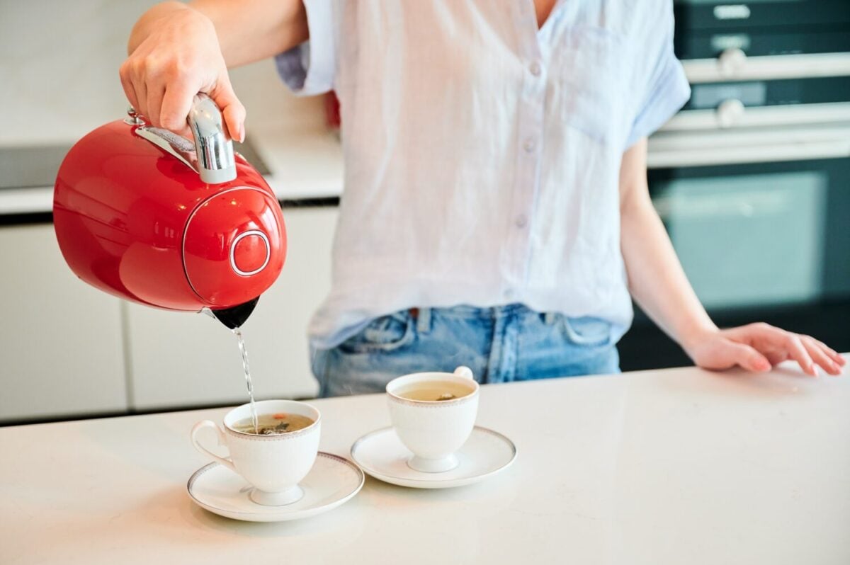 Cropped picture of woman pouring water into cup. Young female making tea for two. Woman holding red kettle over two cups.