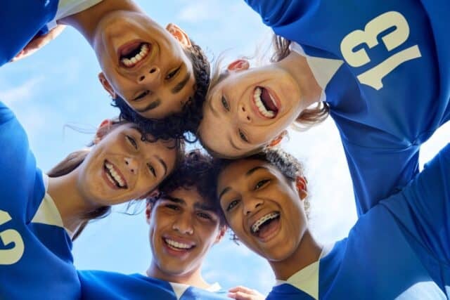 Portrait of young sports team in a close huddle smiling. Group of guys and girls forming a circle with happy faces before match. Cheerful multiethnic teammates looking at camera in unity.