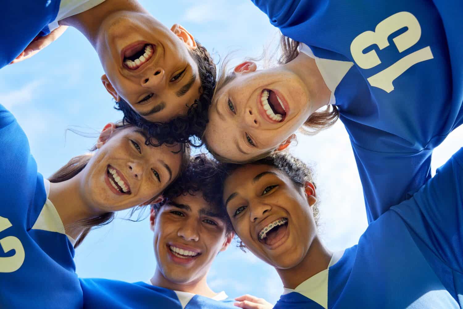 Portrait of young sports team in a close huddle smiling. Group of guys and girls forming a circle with happy faces before match. Cheerful multiethnic teammates looking at camera in unity.