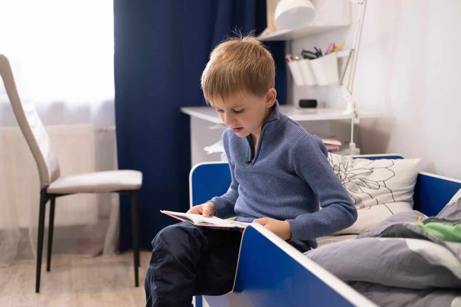 children's book to read. We learn from books. School education. Cute little boy is sitting on the bed in his room and reading book.