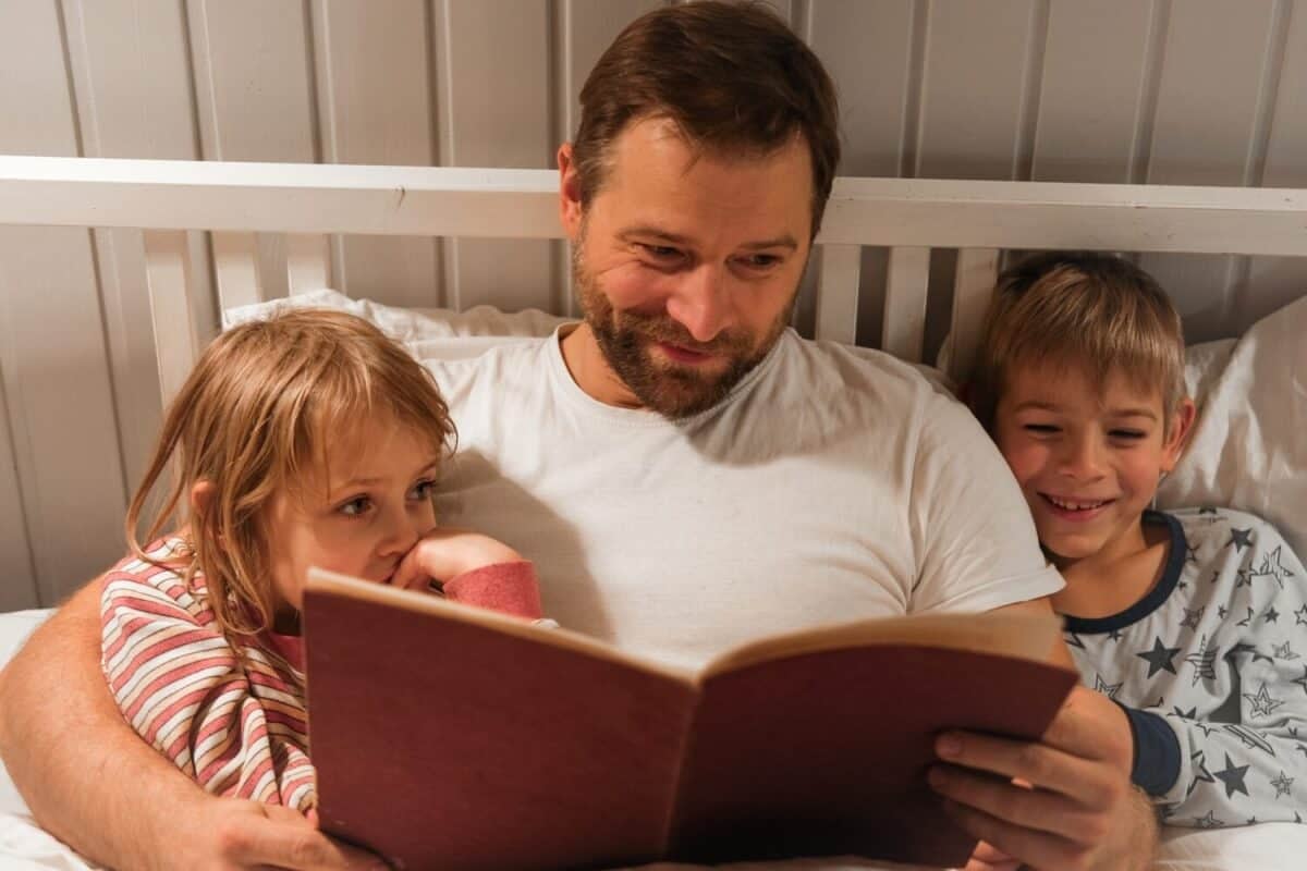 Happy mature 40 year old father reading book to his curious children lying in a bed before the sleep. Dad, little daughter and son spending time together. Family read the fairy story. Evening bedroom.