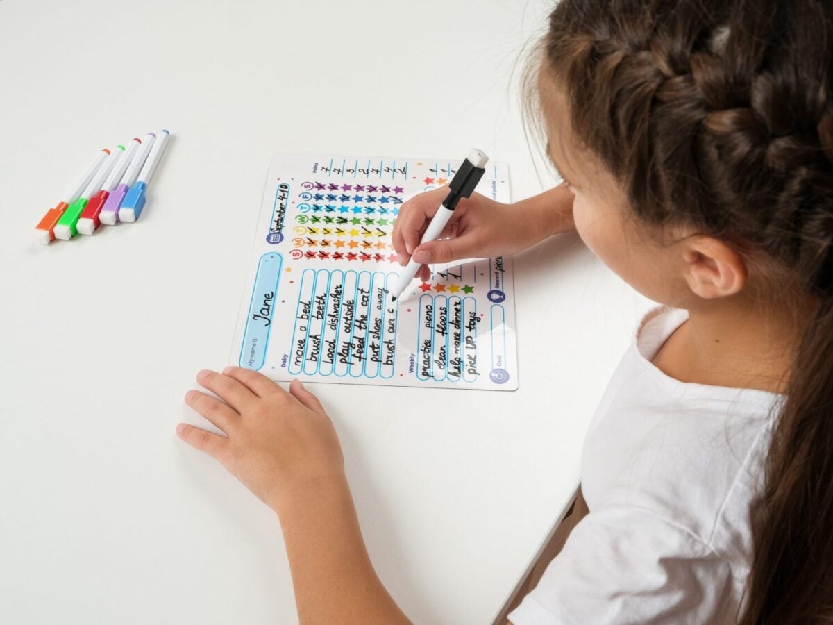 Cheerful school-age girl writes on a reward chart attached to a whiteboard. The system helps children develop discipline, track accomplishments, and improve behavior using visual incentives.