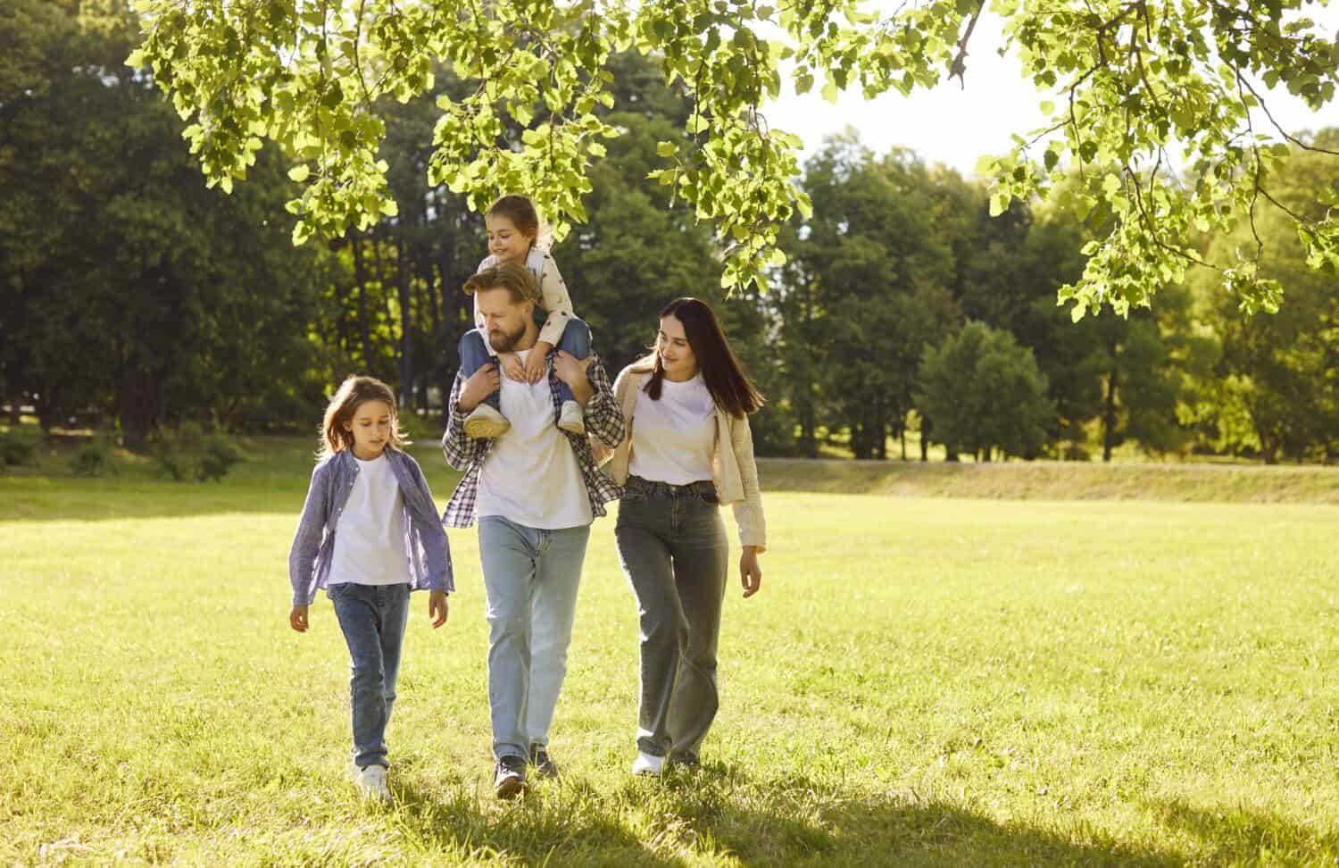 Happy family walking together in park. Mother, father and children enjoy summer or spring green nature and fun healthy walk, dad carrying child on shoulders. Family love, stroll, bonding concept