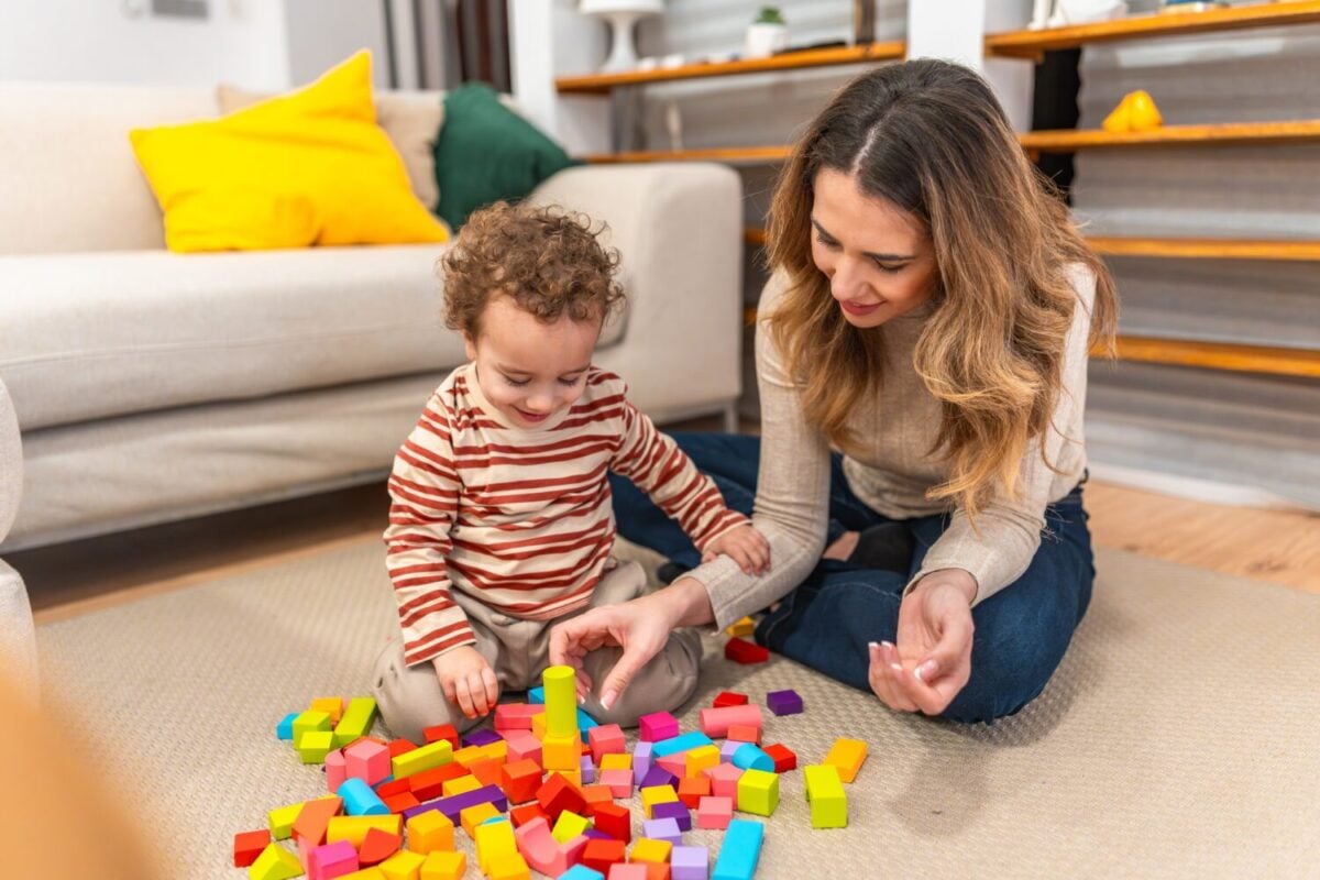 Loving mother and her smiling toddler son are sitting on the floor at home, engaging in creative play with colorful wooden building blocks, fostering developing skills and early childhood learning