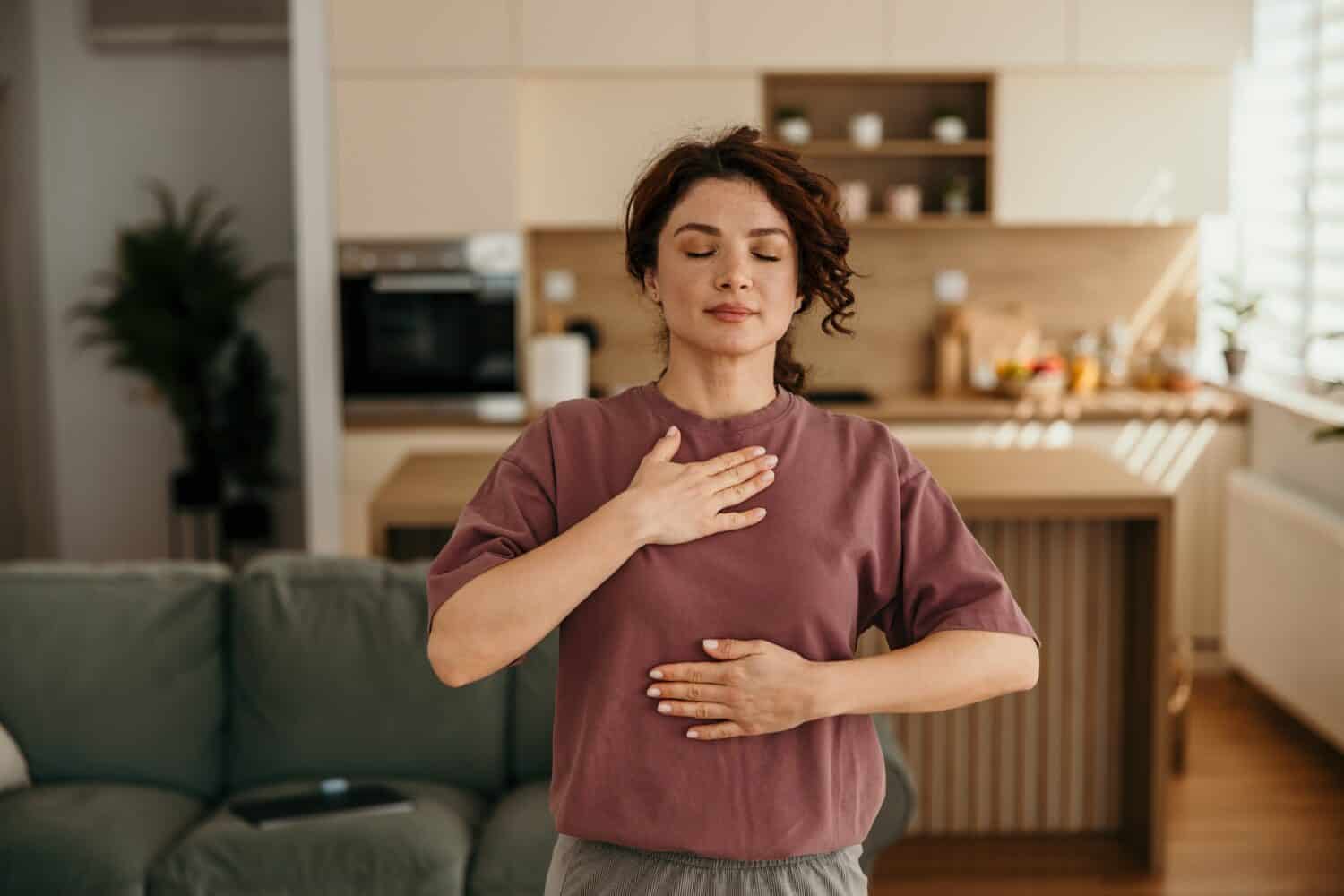 Woman standing with eyes closed, placing hands on chest and stomach, doing breathing exercise