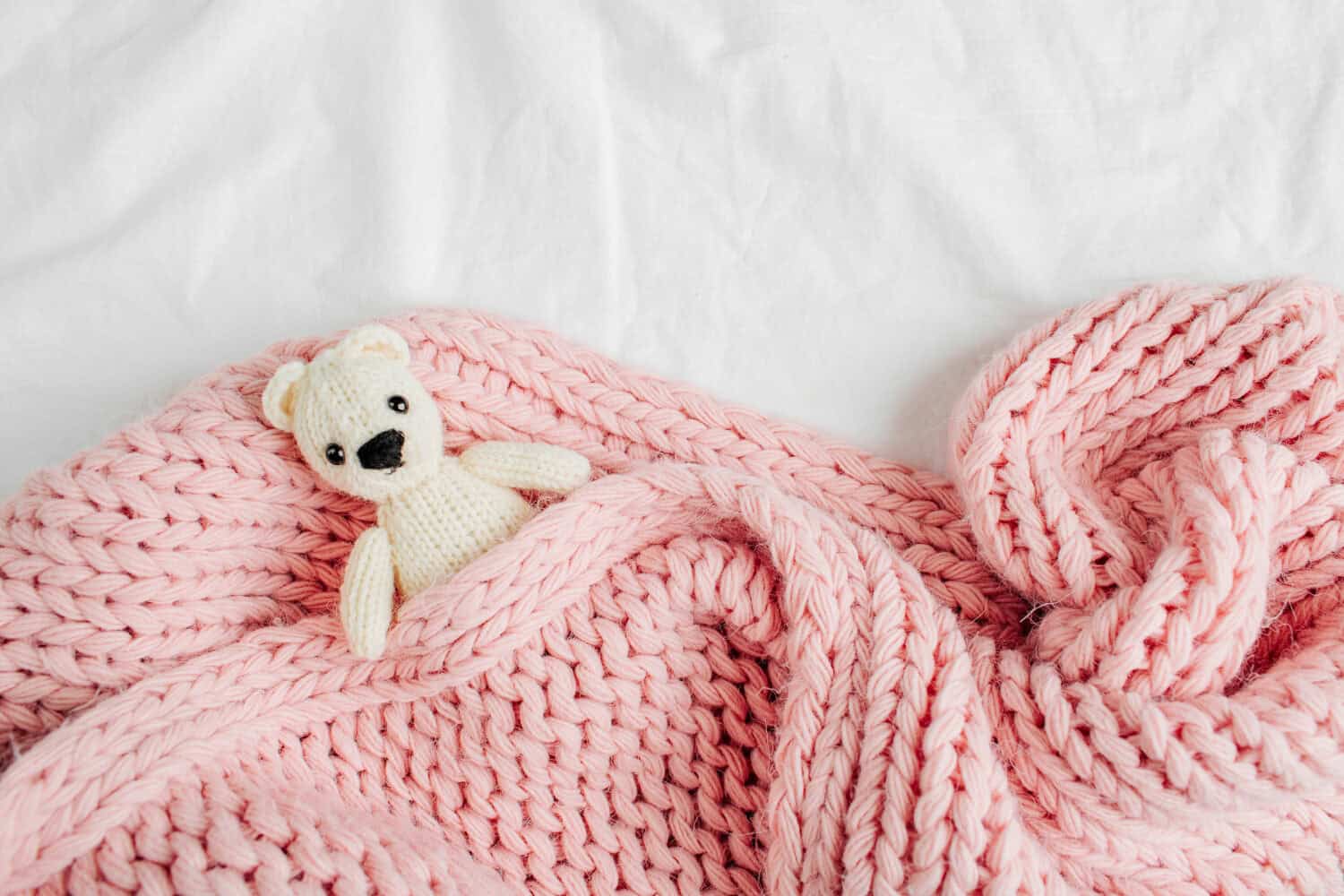 A small knitted baby toy-bear is covered with a warm blanket, flat lay, top view