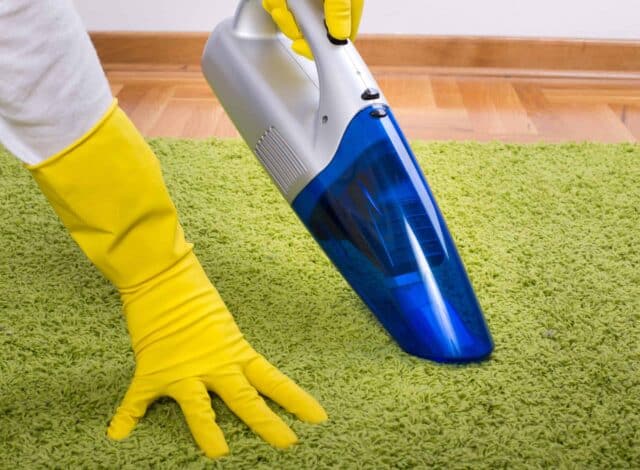 Close up of female hands with rubber gloves holding cordless vacuum cleaner and hoovering green carpet on parquet floor