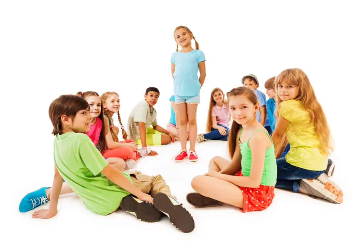 A group of kids sitting in a circle with one kid standing in the middle.