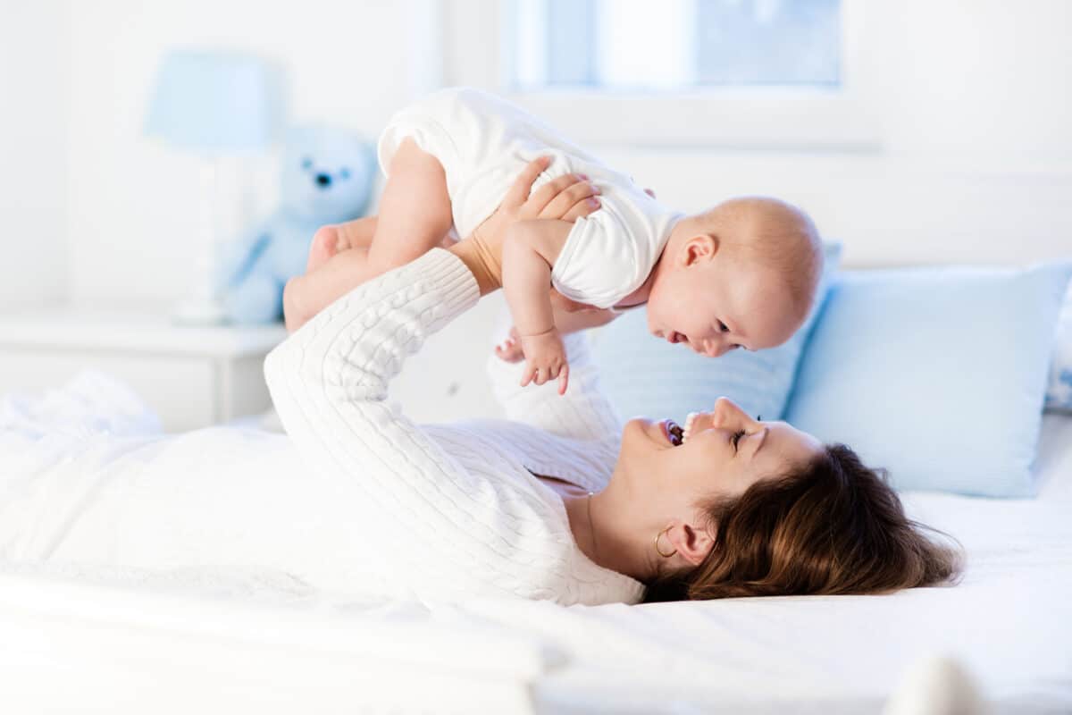 Mother and child on a white bed. Mom and baby boy in diaper playing in sunny bedroom. Parent and little kid relaxing at home. Family having fun together. Bedding and textile for infant nursery