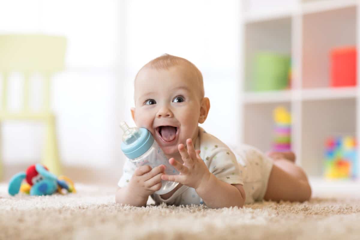 Cute baby boy drinking from a bottle. Kid lying on the carpet in the nursery at home. The smiling child is 7 months old.