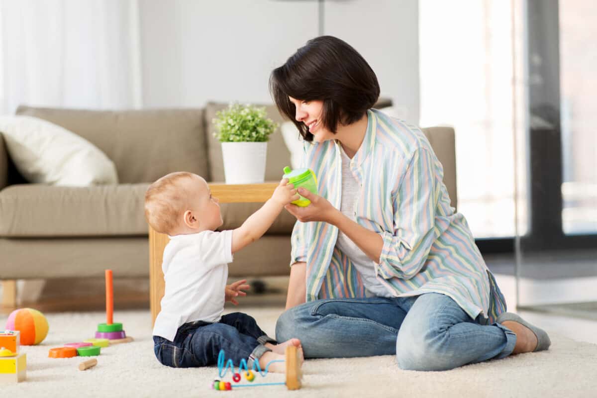 happy mother with little baby son playing with developmental toys and a sippy cup at home