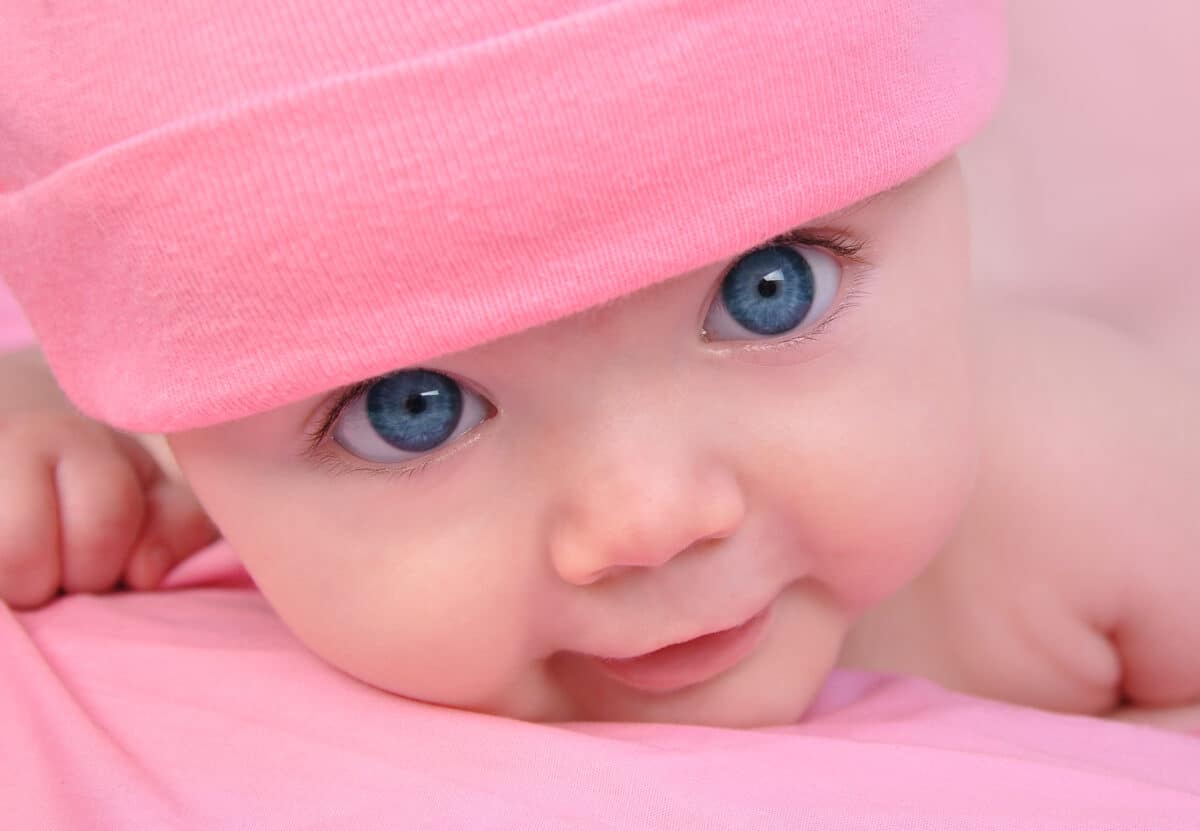 A cute little baby girl is staring up and is on a pink blanket. She is wearing a pink hat and has big blue eyes.