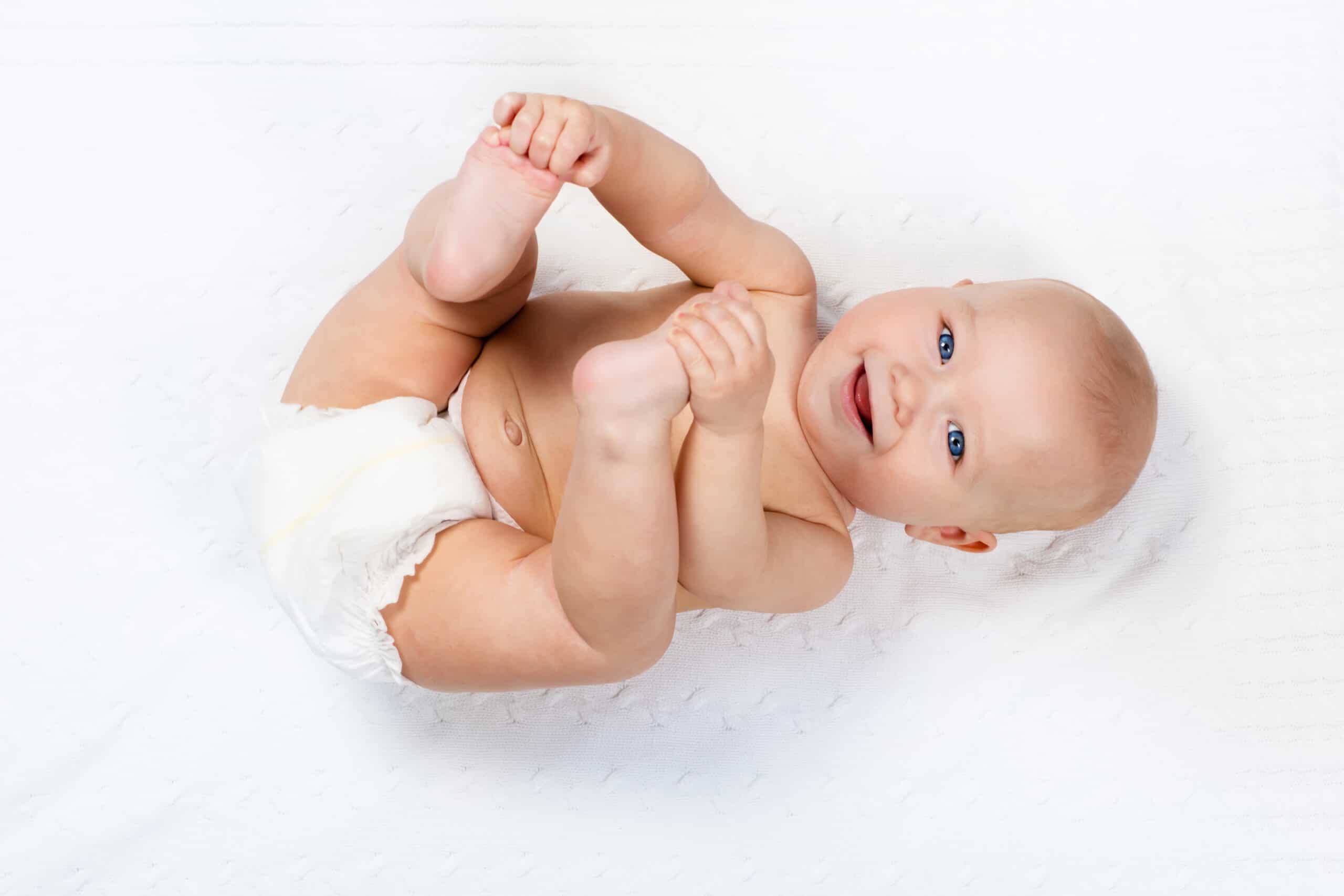 Funny little baby wearing a diaper playing on a white knitted blanket in a sunny nursery.