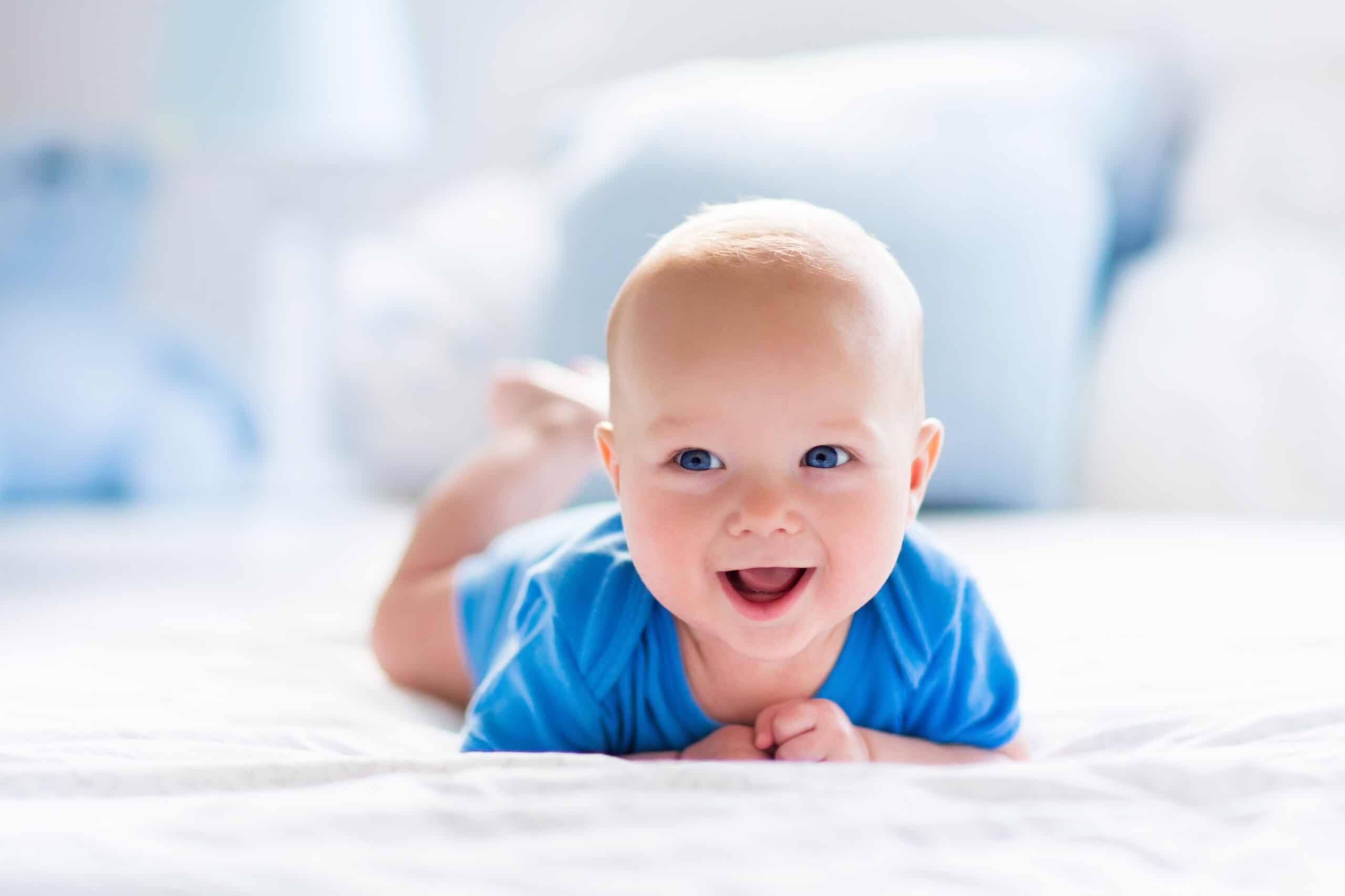 Adorable baby boy in a white sunny bedroom. Newborn child relaxing in bed. Nursery for young children. Textile and bedding for kids. Family morning at home. Newborn kid during tummy time with toy bear.