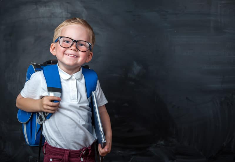 Little boy with glasses holding a backpack in front of a chalkboard.