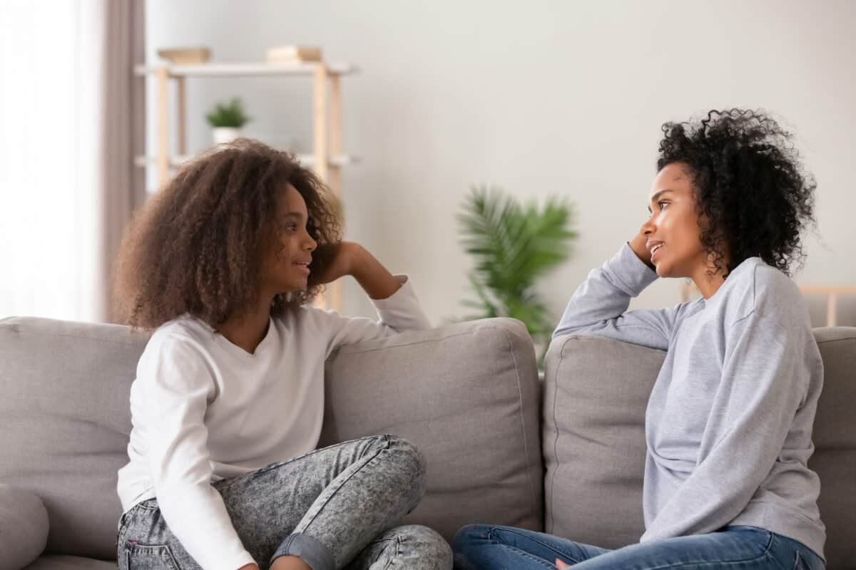 Young african black mother chatting with teen daughter, older sister or friendly nanny and adolescent girl talking sitting together on sofa at home. Good relations different aged people trust concept