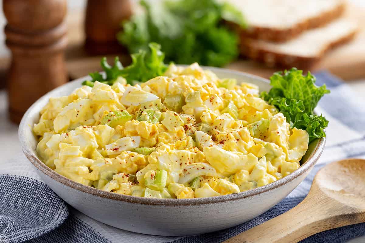 Egg salad in a bowl with bread slices and lettuce on a cutting board in background