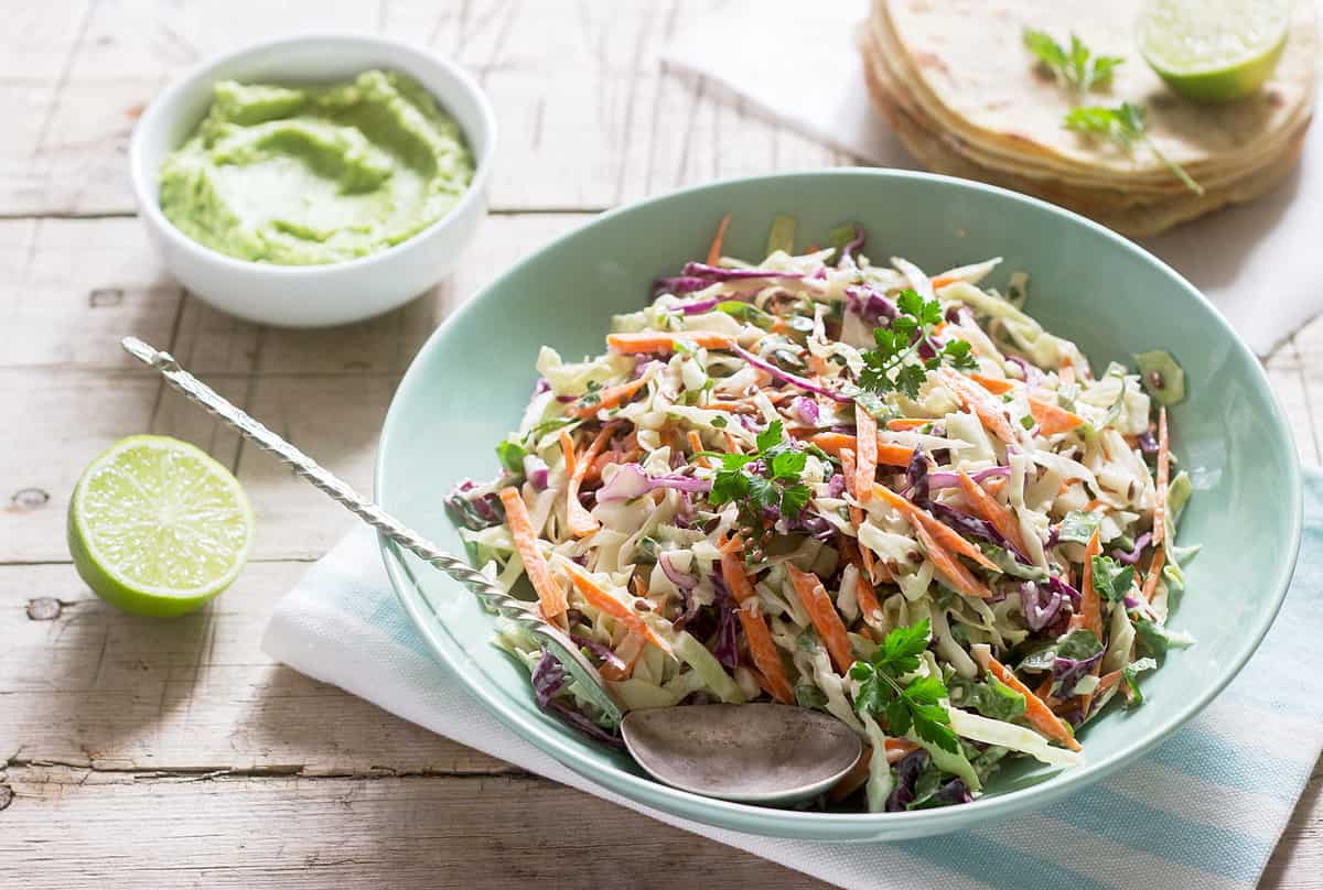 Coleslaw is made from cabbage, carrots, and various herbs, and served with tortillas and guacamole on a wooden background.