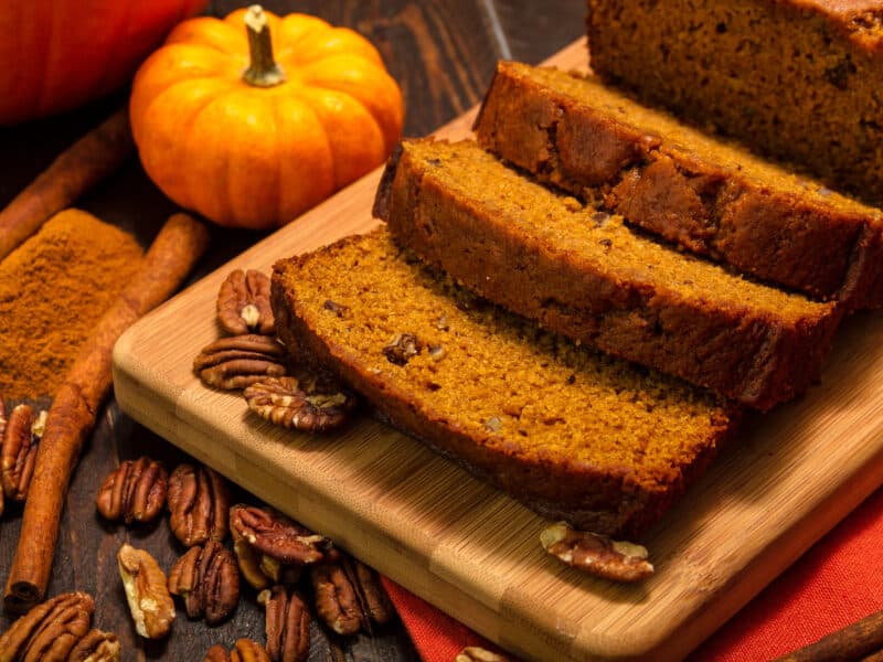 Pumpkin nut bread made with pecans on a cutting board with extra sprinkling of pecans next to a small pumpkin