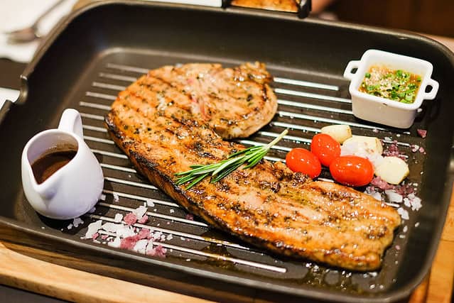 Grilled pork tomahawk (pork chop) with tamarind sauce, black pepper sauce, tomato, garlic, and salt served on a hot black pan.