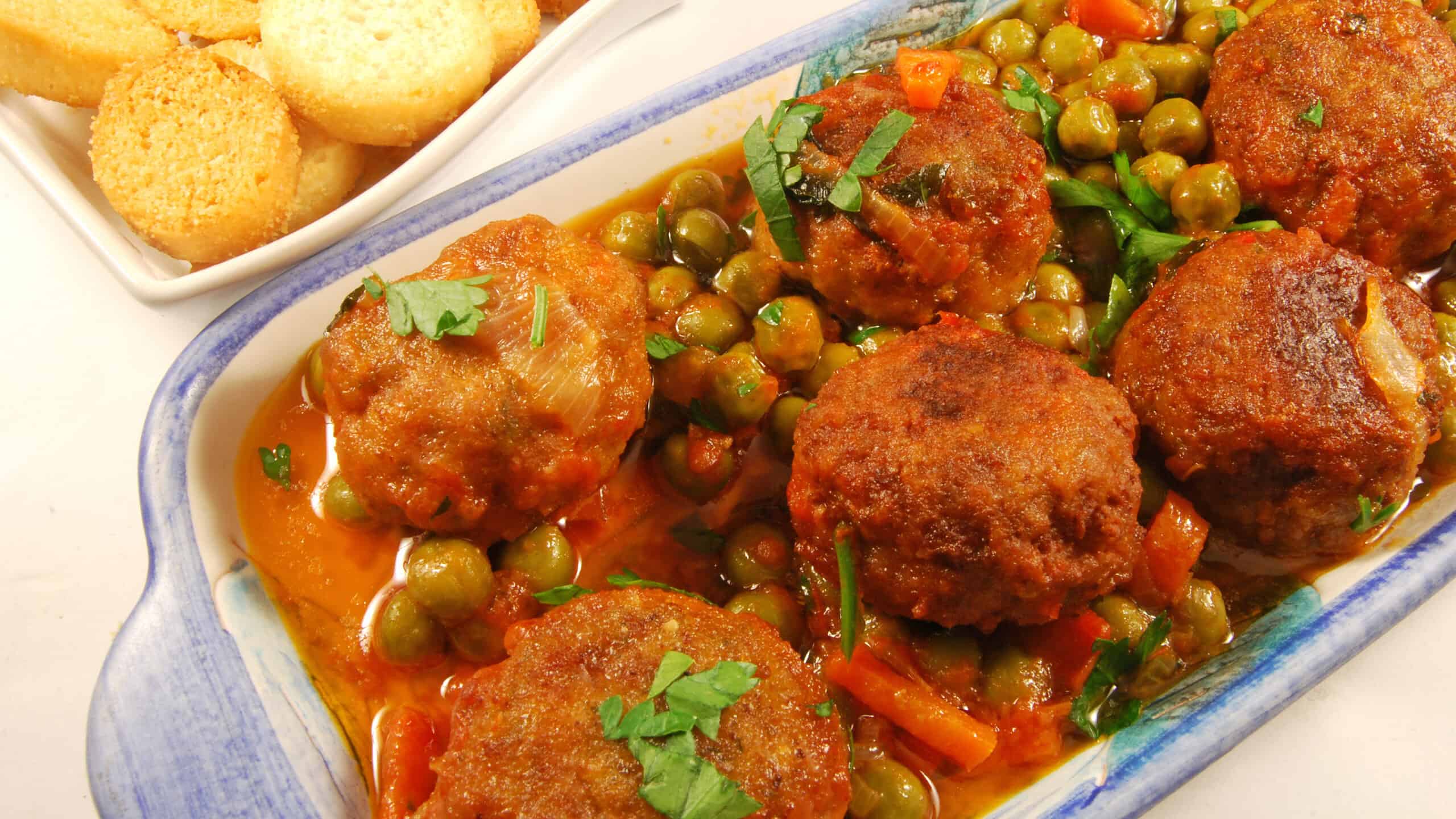 A tray of meatballs in tomato sauce with green peas accompanied by toasted bread with rosemary